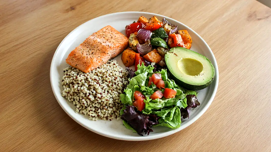 Ausgewogene Mahlzeit mit Lachs, Quinoa, Ofengemüse, Salat und Avocado auf einem hellen Holztisch